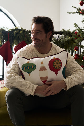 A man in a cream sweater sits on a green couch, hugging the Hanging Ornaments Lumbar Pillow. Behind him are red stockings, garland, and a Christmas tree decorated with red ornaments.
