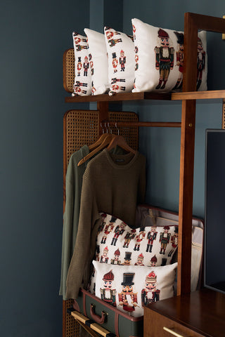 A wooden wardrobe displays hangers with shirts and is decorated for the holidays, featuring the Nutcracker Holiday Lumbar Pillow and a vintage suitcase on the lower shelf.
