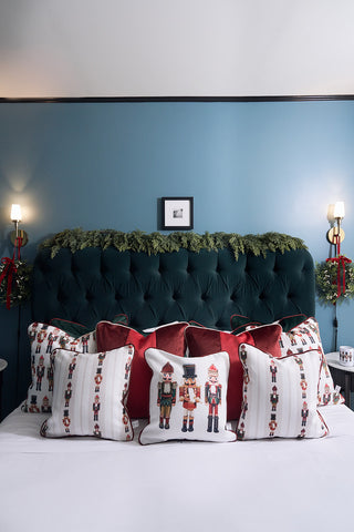 A bed with Nutcracker Holiday Pillows and a dark green tufted headboard adorned with evergreen garland, set against a blue wall featuring wall sconces and wreaths with red ribbon accents on each side.