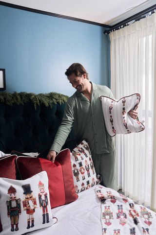 A man in green pajamas stands by a bed with nutcracker-themed bedding, smiling as he arranges a Red Blocking Velvet Pillow. The room has blue walls, white curtains, and festive greenery on the headboard.