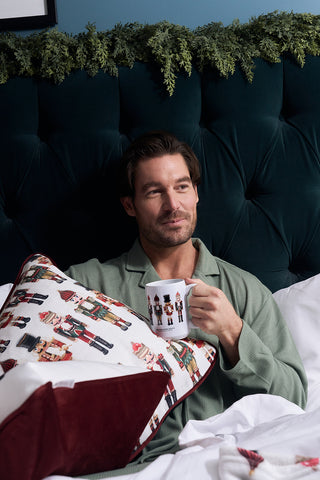 A man in green pajamas sits in bed against a dark, tufted headboard with greenery, holding a festive mug and a Nutcracker Line Up Pillow. He looks relaxed and content.