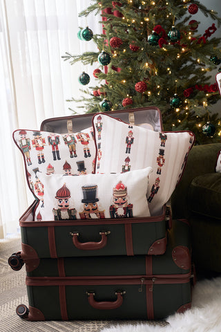 Two vintage suitcases sit open in front of a decorated Christmas tree. The top suitcase is filled with festive Sewing Down South holiday velvet pillows, including the Nutcracker Line Up Pillow. Ornaments and string lights hang on the tree behind.