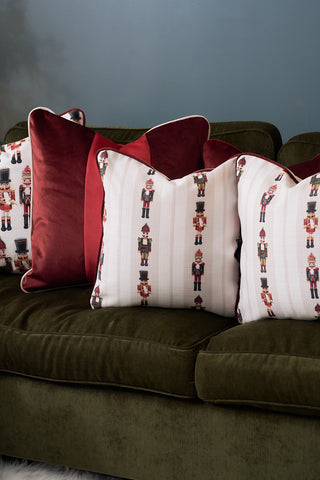 Four pillows are on a dark green sofa: two Red Blocking Velvet Pillows with a velvety finish, and two holiday pillows with white backgrounds and nutcracker soldier patterns. The wall behind is a muted blue shade.