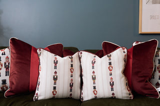 Five decorative pillows rest on a dark sofa against a blue wall: three feature nutcracker holiday designs, while two are Red Blocking Velvet Pillows with white trim. A framed picture is partially visible on the right.
