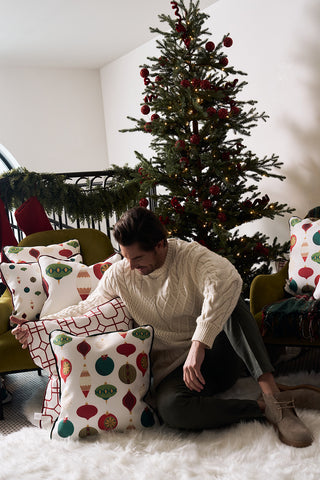 A man in a cream sweater sits on a fluffy white rug, surrounded by festive holiday decor pillows, including the Red Holiday Lattice Oversized Pillow. Behind him, a decorated Christmas tree and garland over the railing complete the cozy scene.