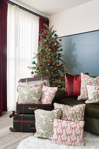 A festive Christmas tree with red and green ornaments stands by sheer-curtained windows; a green sofa decorated with holiday pillows including the Oh Mistletoe Oversized Pillow sits near two stacked suitcases on a white rug.