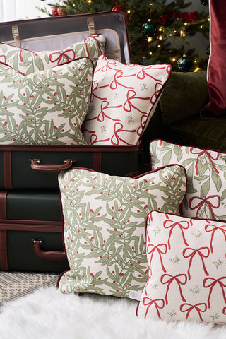 A stack of decorative holiday pillows, featuring the Jolly Holly Pillow with green leaves and a red bow, is arranged on dark suitcases beside a Christmas tree decorated with ornaments.