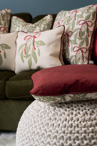 A cozy living room with a green couch adorned with green leaf and red ribbon cushions, the Jolly Holly Pillow for holiday cheer, and a white knitted pouf in the foreground.