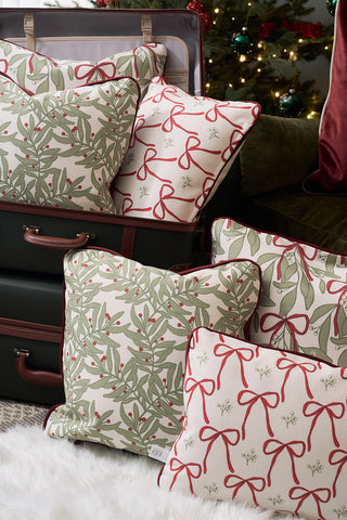 Decorative pillows, including the Jolly Holly Pillow featuring green leaf and red ribbon patterns, are arranged on stacked suitcases with a Christmas tree and holiday lights in the background.