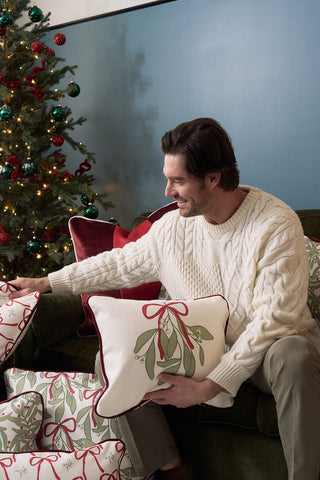 A man in a cream cable-knit sweater sits on a green couch, smiling and holding the Bows & Mistletoe Lumbar Pillow. Holiday pillows surround him, with a glowing tree adorned with lights and red ornaments in the background.