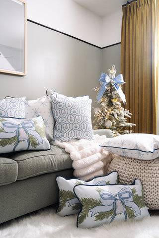 A cozy living room corner features a green sofa with the Holiday Swag Extended Lumbar Pillow, complemented by winter-themed blankets. Nearby, a small decorated Christmas tree with a blue bow stands beside gold curtains on a soft white rug.