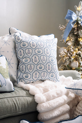 A cozy living room with a light blue couch, patterned pillows including the Silver Bells Pillow, a white fluffy blanket, and a decorated Christmas tree featuring ornaments and a large blue ribbon bow in the background.