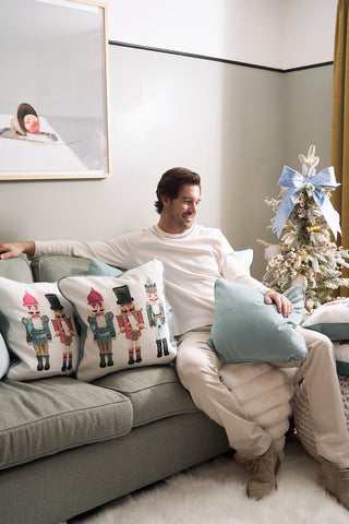 A man in light-colored clothes sits on a sofa holding a blue pillow, next to the Pastel Nutcracker Pillow from Sewing Down South and other holiday pillows. Behind him is a small Christmas tree with white ornaments and a blue bow, plus a framed print on the wall.