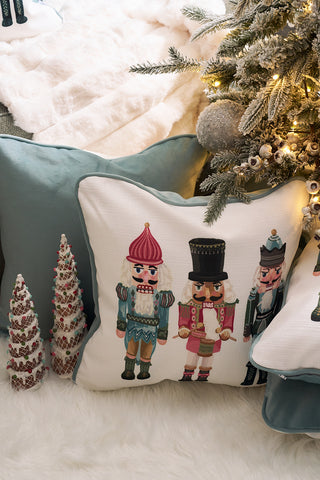 A Pastel Nutcracker Pillow adds a festive touch next to a Christmas tree and ornaments on a soft white rug, creating a cozy holiday scene.