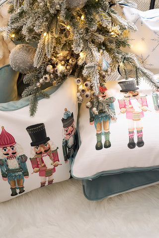 A charming Pastel Nutcracker Pillow from Sewing Down South sits on a white furry rug under a Christmas tree with snow-dusted branches, sparkling ornaments, and twinkling lights.