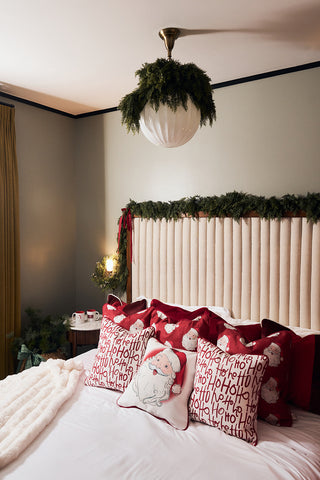 A cozy Christmas bedroom with a cream headboard, velvet holiday accents, red decor, garlands above the bed, a white fur blanket, a greenery-adorned ceiling light, and a Vintage Repeating Santa Pillow for festive charm.