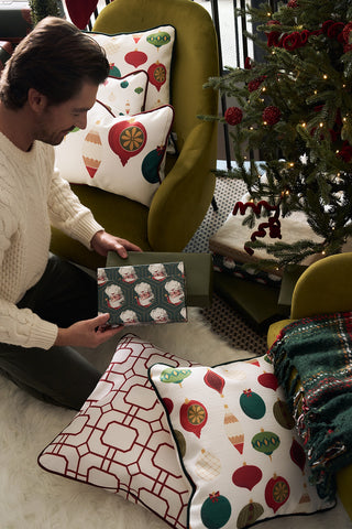 A man in a cream sweater sits on a white rug by a decorated Christmas tree, holding a Santa-themed gift. The Classic Ornaments Pillow and folded blankets on green chairs nearby create a cozy, festive atmosphere.