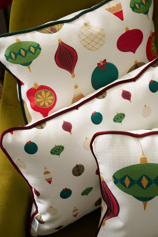 Three accent pillows with festive holiday ornament patterns in red, green, and gold are stacked on an olive green chair. The set features the Hanging Ornaments Lumbar Pillow for a modern seasonal touch.