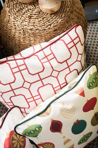 Two decorative pillows are stacked on a woven basket. The top pillow is the Red Holiday Lattice Oversized Pillow, while the bottom is a colorful holiday decor pillow. Both sit on a patterned rug, bringing festive charm to your space.