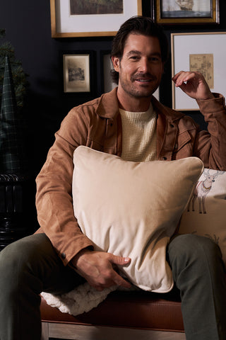 A man with medium-length brown hair and a trimmed beard sits on a sofa, smiling and holding the Holiday Reindeer Pillow. He wears a tan jacket, cream sweater, and olive-green pants. Framed art decorates the dark wall behind him.