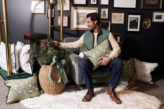 A man in a green vest sits on a gray chair in a cozy room, holding an accent pillow. Nearby are more pillows, including the Winter Sports Gear Lumbar Pillow, plus a basket with pine branches and framed art for a festive atmosphere.
