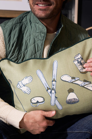A man in a green quilted vest and cream sweater holds the Winter Sports Gear Lumbar Pillow, decorated with ski equipment illustrations—perfect for adding a touch of Southern decor to any space.