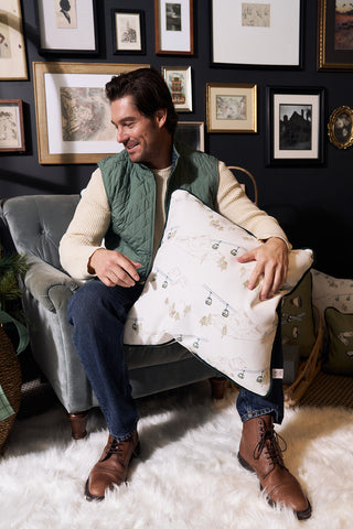 A smiling man in boots and a green quilted vest sits on a green armchair, holding a Ski Resort Pillow with a winter motif, surrounded by framed art on a dark wall and a fluffy white rug underfoot.