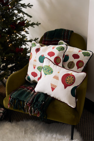 A green armchair with a dark green plaid blanket and two festive accent pillows, including a Hanging Ornaments Lumbar Pillow, sits next to a decorated Christmas tree adorned with sparkling lights.