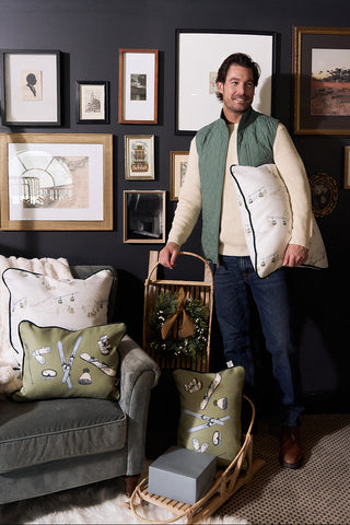 A man stands by a gray armchair holding the Winter Sports Gear Lumbar Pillow. He wears a cream sweater, green vest, and jeans. The room features framed art on dark walls, modern decor, baskets, and a small wreath.