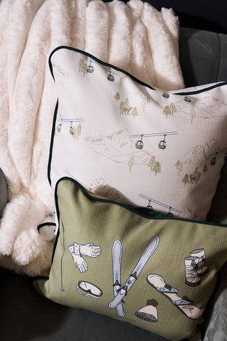 Two decorative pillows on a plush white blanket: one is the Winter Sports Gear Lumbar Pillow with vintage ski gear illustrations on olive-green, the other features a ski lift and mountain print—perfect après-ski home decor.