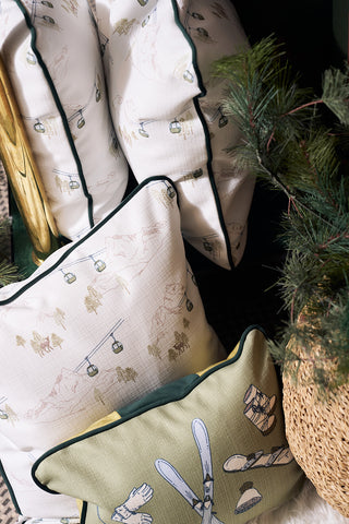 A close-up of the Ski Resort Pillow featuring ski lift, mountain, and equipment illustrations, stacked by a pine branch and woven basket, creating a cozy vintage ski decor and charming holiday vibe.