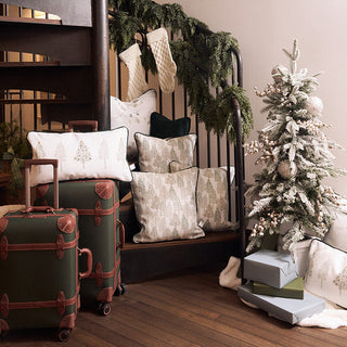 A cozy holiday scene with a small decorated Christmas tree, wrapped gifts, stacked pillows with tree patterns, green suitcases, and stockings hanging on a garlanded spiral staircase.