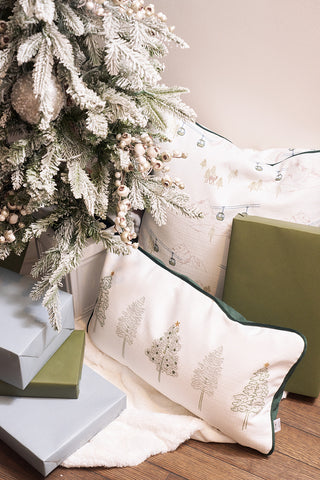 A Ski Resort Pillow with tree designs sits next to a frosted Christmas tree with white berries, stacked green and gray gift boxes, and a fluffy white rug on a wooden floor.