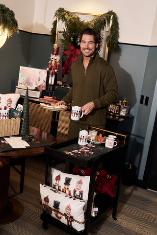 A smiling man stands behind a cart decorated with holiday-themed items, like the Nutcracker Holiday Lumbar Pillow, mugs, and treats, in a cozy room adorned with garland and festive holiday decorations.