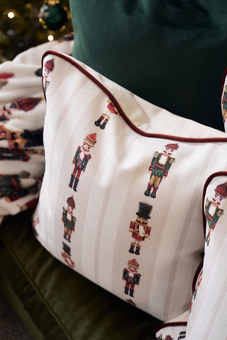 The Neutral Nutcrackers Pillow, featuring small embroidered nutcrackers in rows, rests on a green velvet couch beside a matching blanket. Blurred Christmas decorations in the background enhance the festive holiday pillow display.