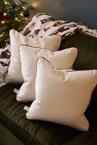 A Neutral Nutcrackers Pillow is placed on a dark green couch alongside white velvet pillows. A festive nutcracker-patterned blanket and a lit Christmas tree add a seasonal touch in the background.