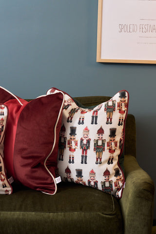 Two decorative pillows on a green armchair: one is a burgundy velvet pillow with white piping, the other is the Nutcracker Line Up Pillow. A framed Spoleto Festival poster hangs on the blue wall behind them.