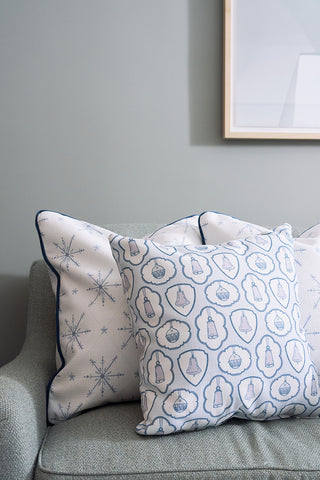 The Silver Bells Pillow, a twill woven holiday accent with blue and white snowflakes, and a nautical-themed pillow with bottles are arranged on a light grey sofa set against a pale green wall.