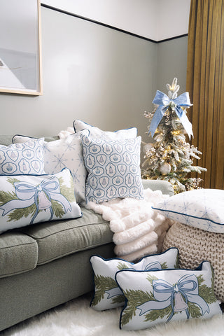 A cozy living room corner features a grey couch with the Luxe Snowflakes Oversized Pillow, a blue Christmas pillow, knit throw, and a small Christmas tree adorned with a blue bow, gold ornaments, and snowy branches.
