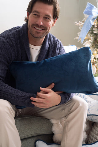 A smiling man with brown hair in a navy sweater and beige pants sits on a couch holding the Holiday Swag Extended Lumbar Pillow. A decorated Christmas tree with a blue ribbon is visible in the background.