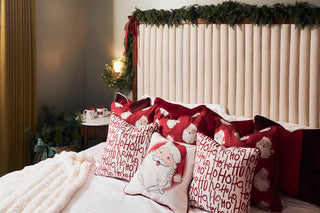 A cozy bed with red and white Christmas-themed pillows, including Santa and Ho Ho Ho designs, sits against a headboard decorated with green garland. Two holiday mugs rest on a nearby nightstand.