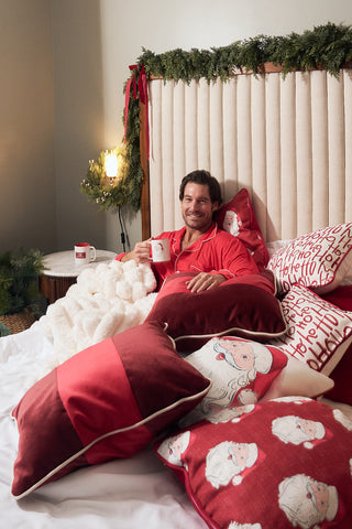 A man in red pajamas sits in bed, smiling and holding a mug, surrounded by red and white holiday pillows, including a Red Blocking Velvet Pillow. Garland on the headboard adds to the festive and cozy atmosphere.