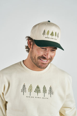 A man wearing a cream sweatshirt and the Christmas Tree Farm Embroidered Hat, both with green tree embroidery and "Sewing Down South," smiles while looking down against a plain light background.