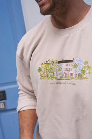 A man in a cozy Holiday Rainbow Row Crewneck Sweatshirt, featuring colorful Charleston, South Carolina houses and the text “charleston, south carolina,” stands by a blue door.