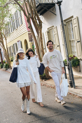 Three people in light outfits walk down a vibrant street with colorful houses and trees, smiling with shopping bags. One wears the cozy Holiday Rainbow Row Crewneck Sweatshirt.