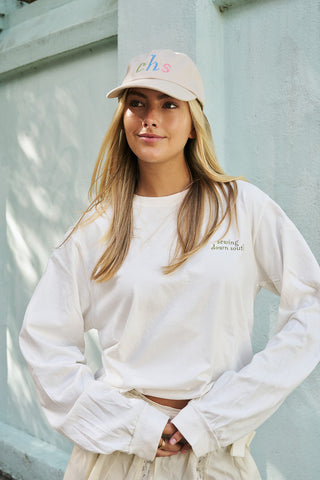 A young woman with long blonde hair stands outdoors, smiling slightly as she wears the Rainbow Row Dad Cap—a beige hat featuring colorful “chs” letters—and a white “going down south” long-sleeve shirt.