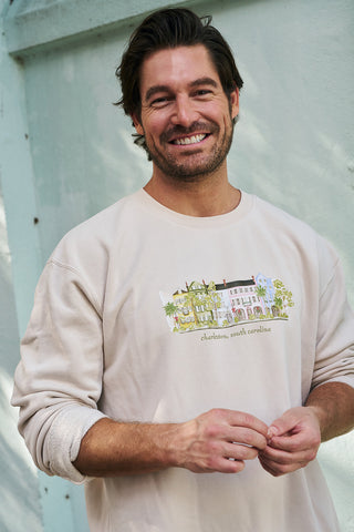 A smiling man with dark hair and a trimmed beard wears the Holiday Rainbow Row Crewneck Sweatshirt, featuring an illustrated Charleston building and text, while standing against a pale green wall.