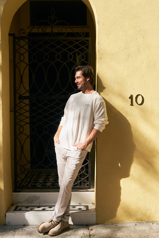 A man wearing a Holiday Rainbow Row Long Sleeve Tee and light pants stands casually in front of a yellow wall with the number 10, leaning against a black wrought iron gate and smiling in the sunlight.