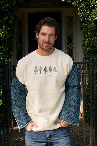 A man stands outdoors by a gate and greenery, wearing the Christmas Tree Farm Embroidered Crewneck featuring tree illustrations and "Driving Down South." His blue jacket is draped off his shoulders, and his hands are in its pockets.