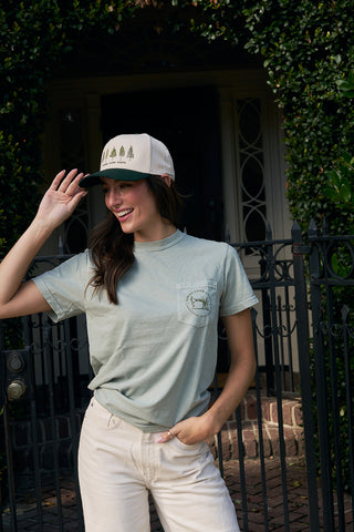 A smiling woman stands outdoors near greenery, wearing the Christmas Tree Farm Tee, beige pants, and a light cap with tree graphics. She holds the brim of her cap with one hand in front of a wrought iron gate.